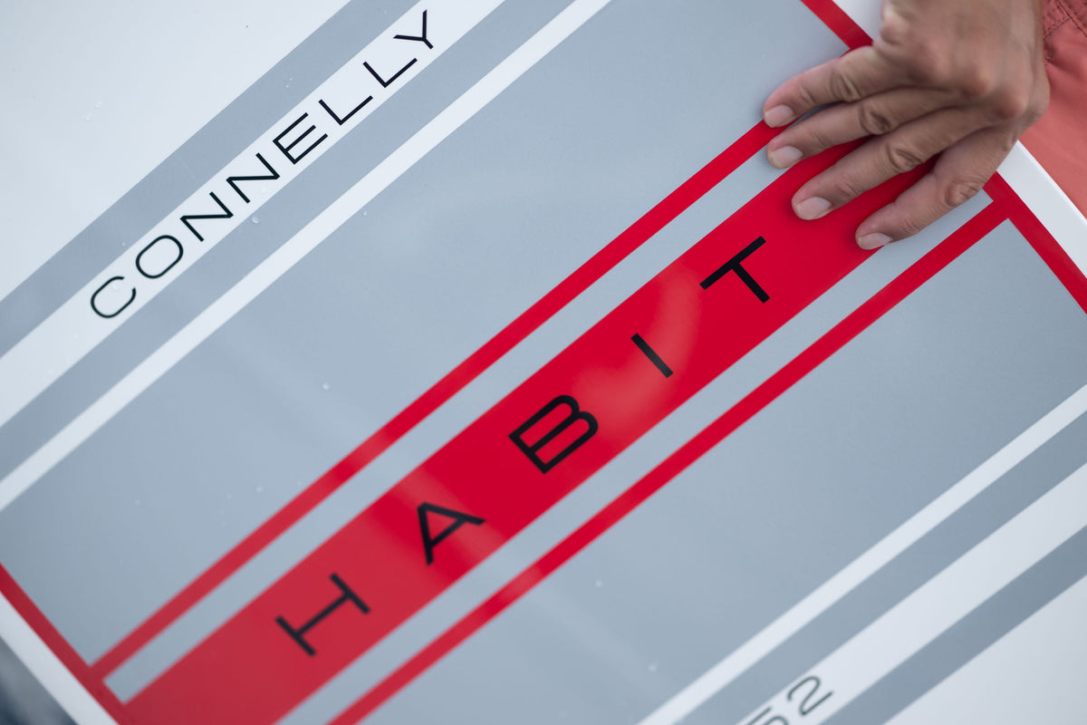 Close-up of the HABIT Skim logo on the board with red and gray stripes and a hand resting on the surface.