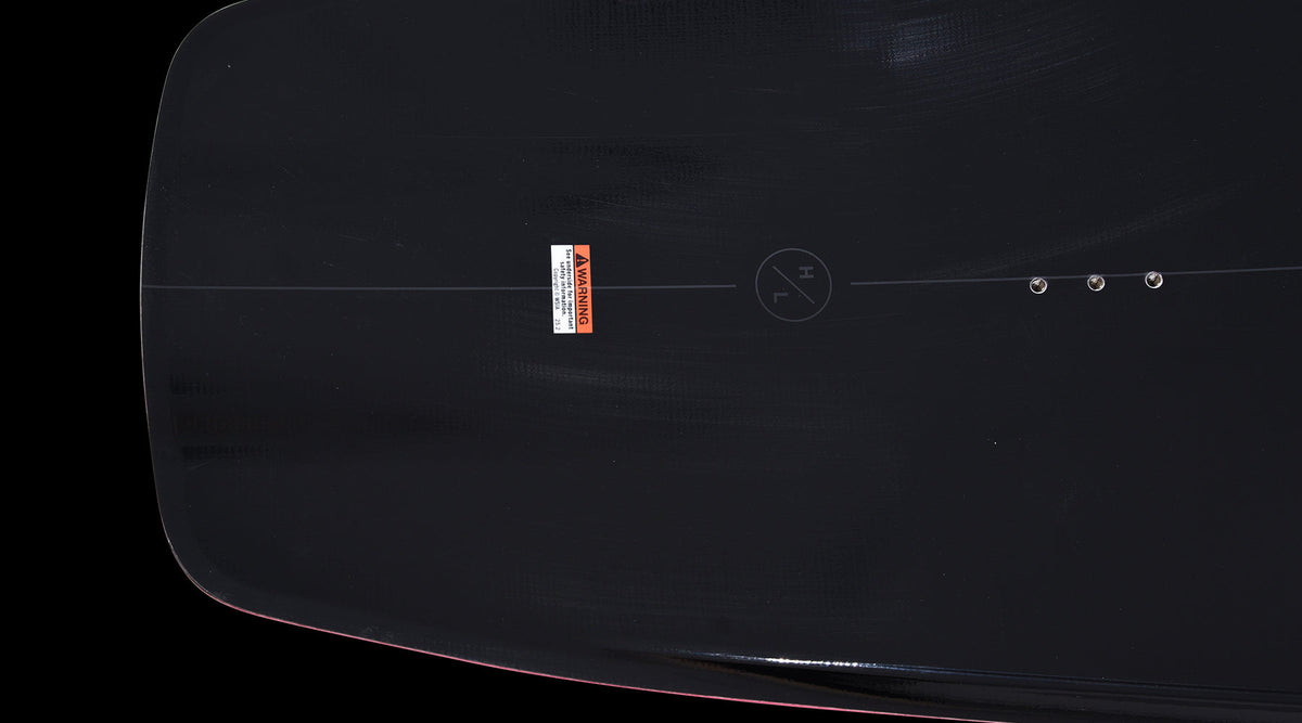 Close-up of mounting inserts and warning label on the black deck of the Pleasure wakeboard.