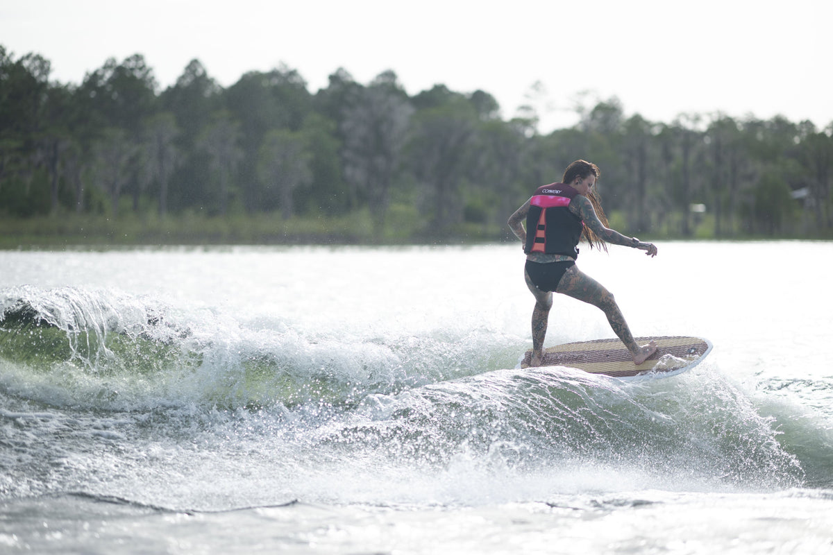 A woman in a pink life vest rides the Lil Easy wakesurf board on a wave, carving through the water.