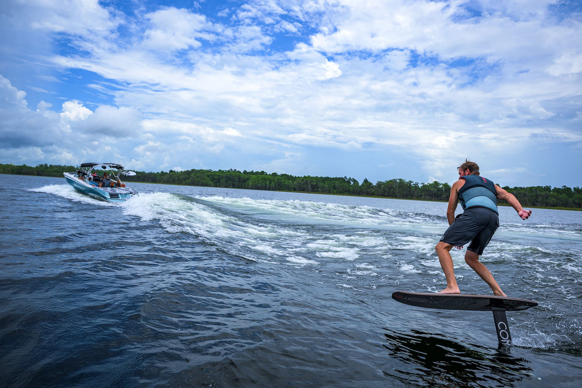 Person on a wakefoil behind a boat on a lake with trees in the background.