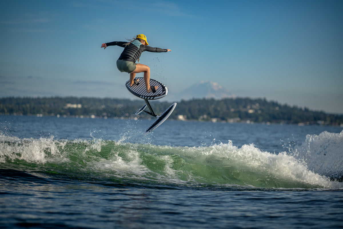 Person gliding on a Majik Karpet over waves, with mountains and trees in the background.