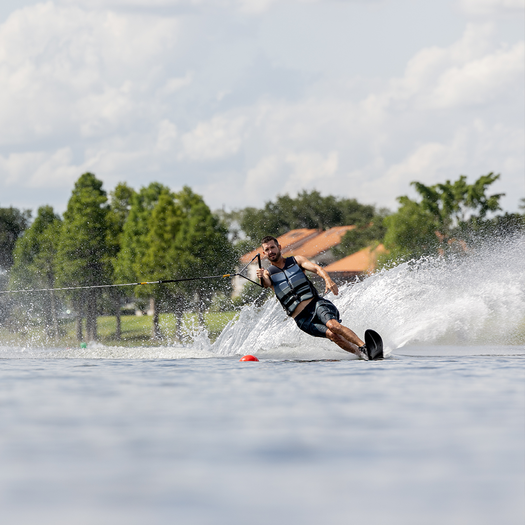 A male water skier in black shorts and a gray vest carves across the water on the Shortline ski.